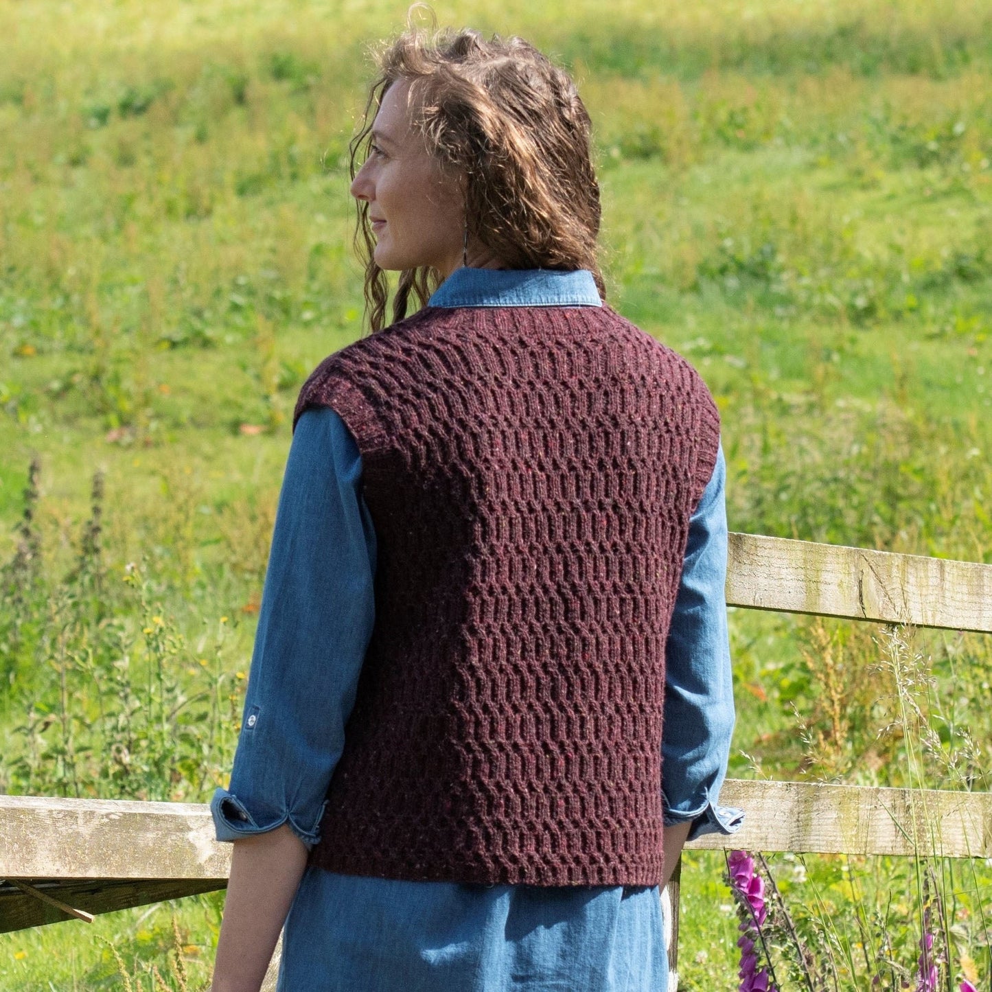 A woman stands in a grassy field with her back to the camera. She wears a handknit sleeveless sweater with an all-over texture stitch on the back.